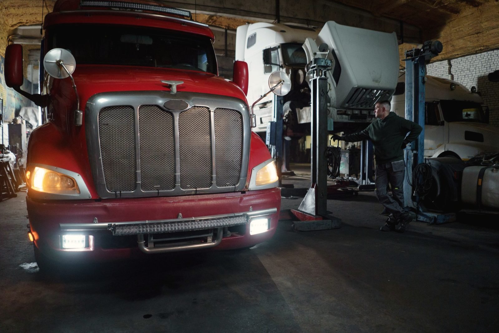 A red tractor with its lights on inside a dim repair shop, other trucks on lifts behind it