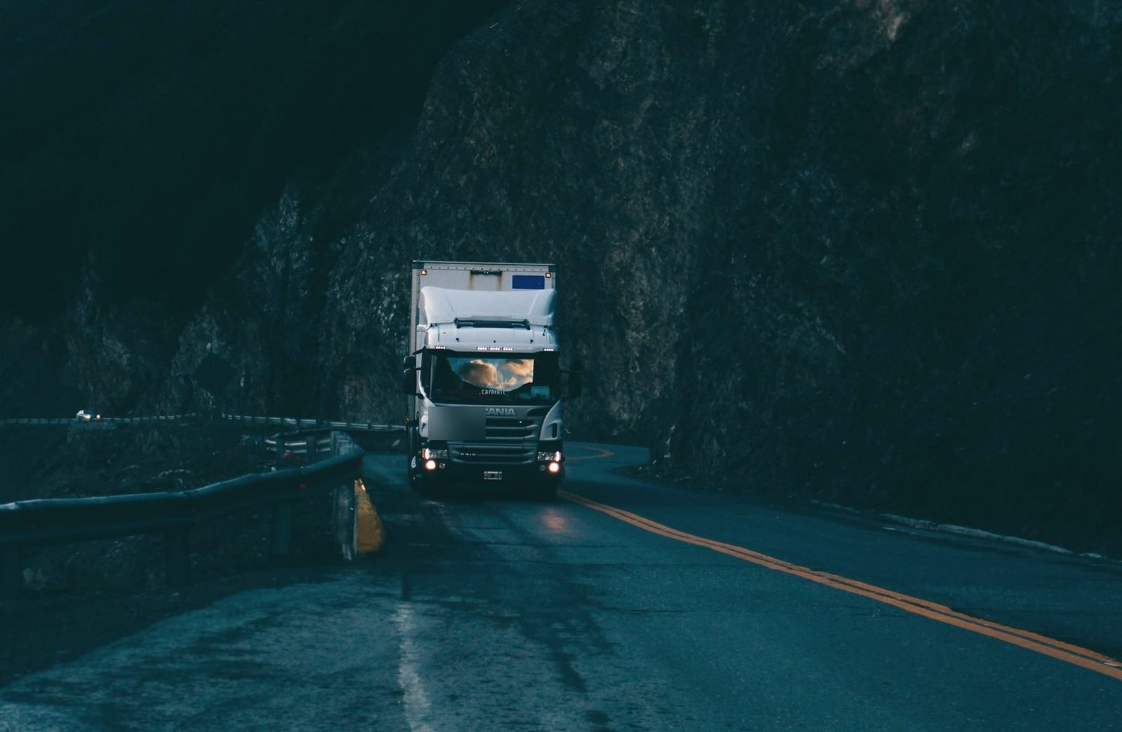 A box truck with headlights on, coming round a dark rock cut at night